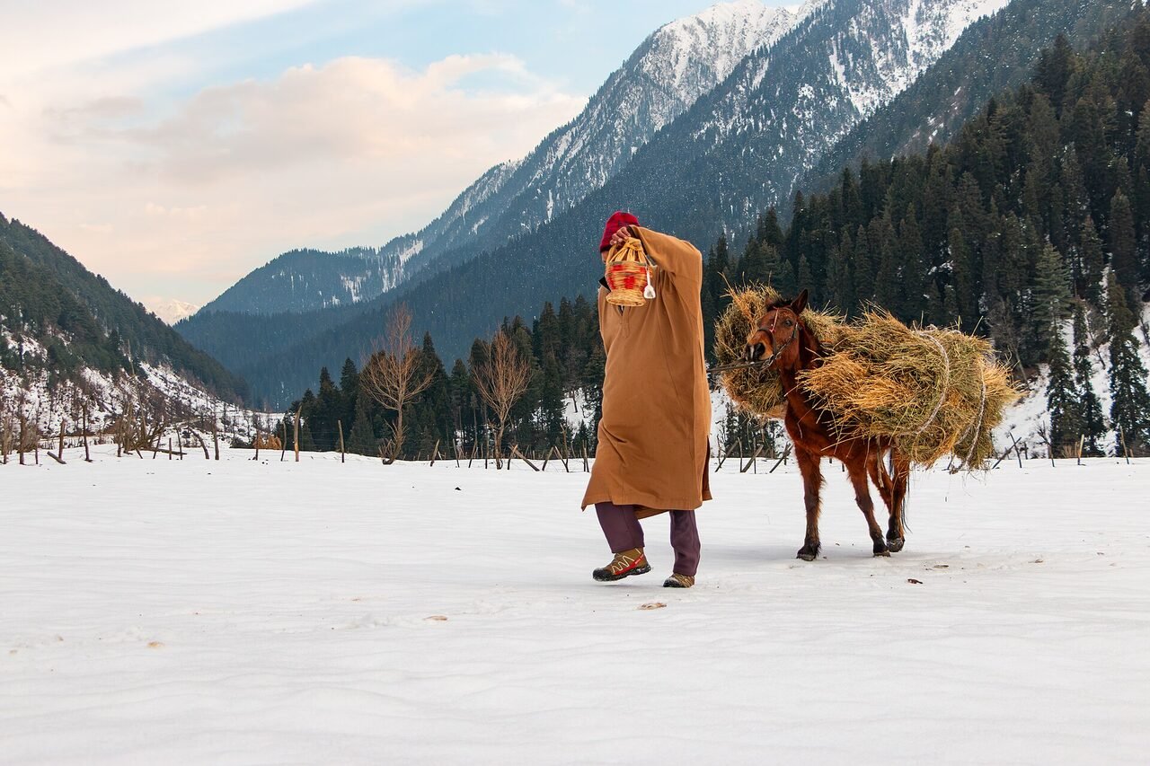 A person in a thick pheran holding a kangri firepot, standing in snow at Aru